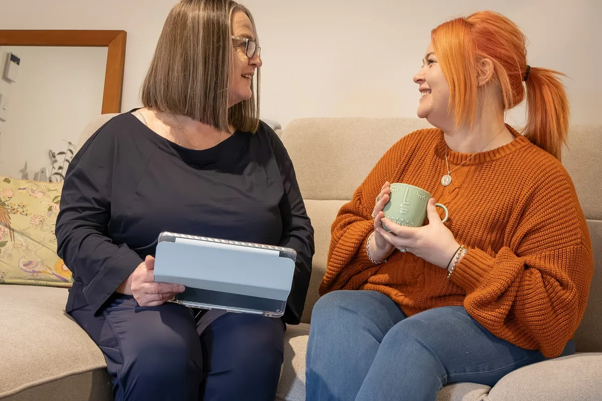 Michelle and a new mother laughing together over a cup of tea on the couch