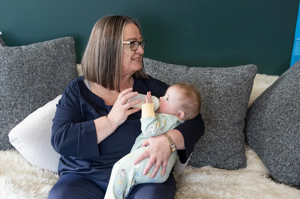 Michelle smiling down at a baby she is bottle-feeding on the sofa in the afternoon